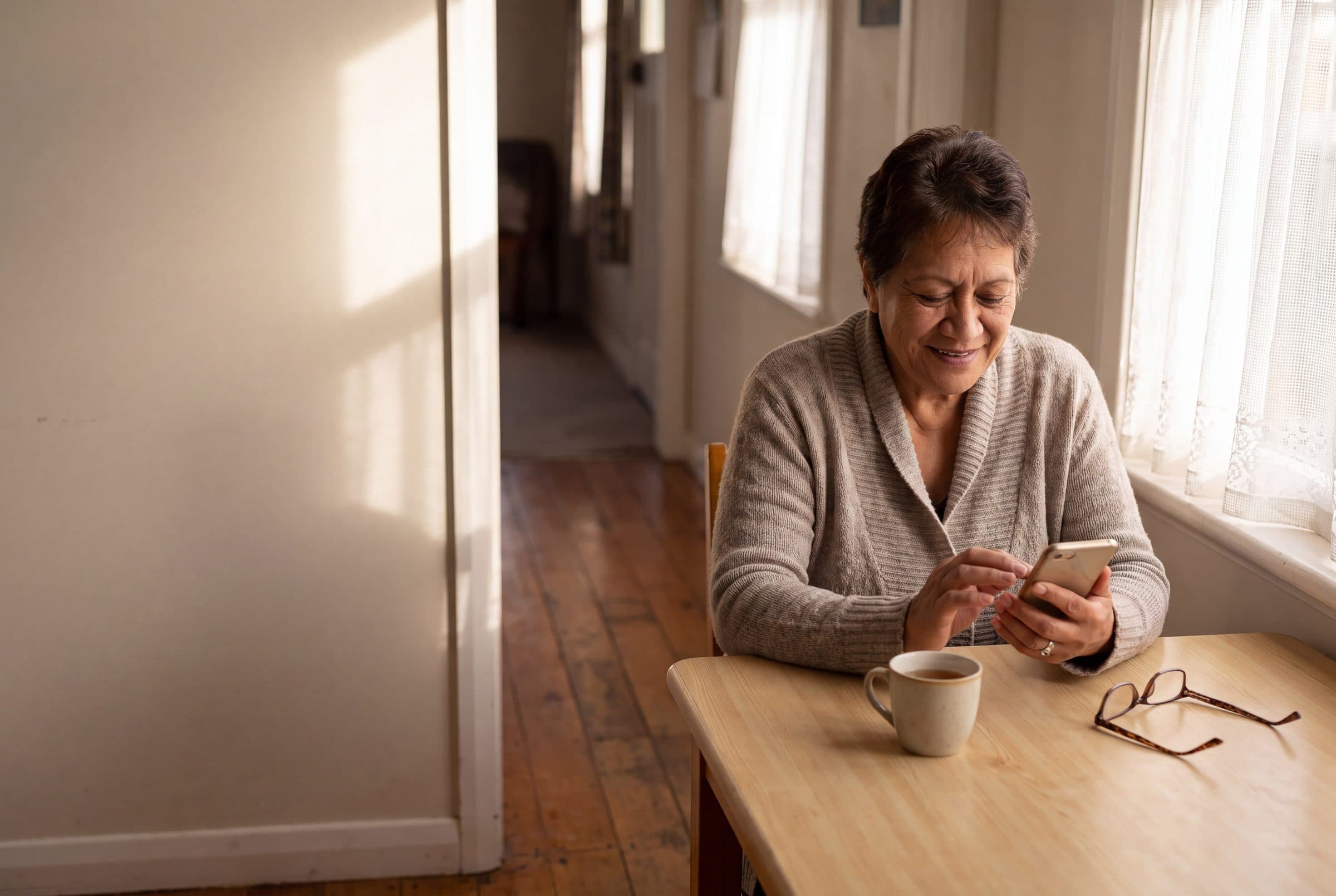 An older woman reading her phone at a sunlit kitchen table